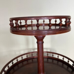 May include: A two-tiered wooden serving stand with a round top and bottom. The top tier has a decorative railing with turned wooden spindles. The stand is made of dark wood.