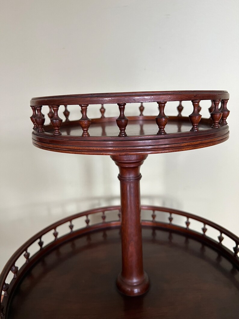 May include: A two-tiered wooden serving stand with a round top and bottom. The top tier has a decorative railing with turned wooden spindles. The stand is made of dark wood.