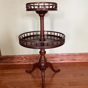 May include: A two-tiered wooden table with a round top and a decorative, scalloped edge. The table has a dark brown finish and stands on three legs.