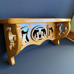 May include: A wooden shelf with a light brown finish. The shelf features decorative cutouts of a steer, cactus, and other shapes. The shelf is supported by two curved brackets and is designed to be mounted on a wall.