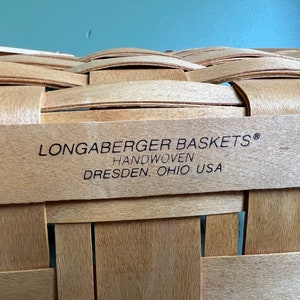 Puede incluir: Primer plano de una cesta de mimbre tejida a mano con una etiqueta que dice "Longaberger Baskets&reg; Handwoven Dresden, Ohio USA".