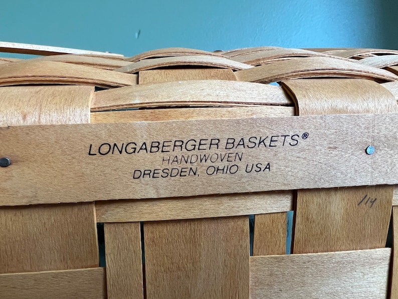 Puede incluir: Primer plano de una cesta de mimbre tejida a mano con una etiqueta que dice "Longaberger Baskets&reg; Handwoven Dresden, Ohio USA".