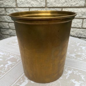 May include: A gold metal cylindrical vase with a slightly flared top rim. The vase has a worn, aged appearance.