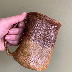 May include: A brown and tan ceramic mug with a textured surface. The mug has a wide, rounded base and a handle.