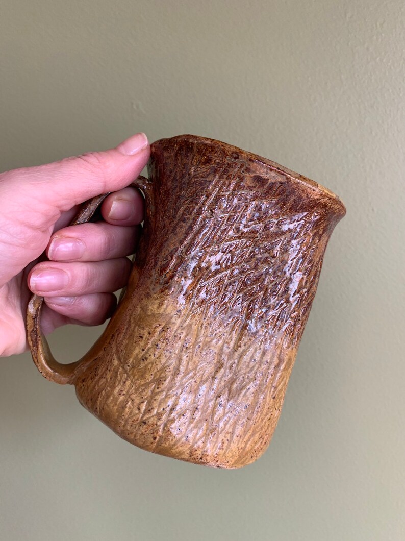 May include: A brown and tan ceramic mug with a textured surface. The mug has a wide, rounded base and a handle.