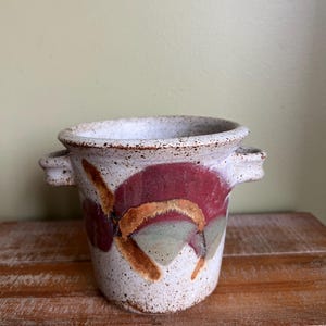 May include: A small, white ceramic planter with two handles. The planter has a speckled glaze and is decorated with abstract brown, red, and green designs.