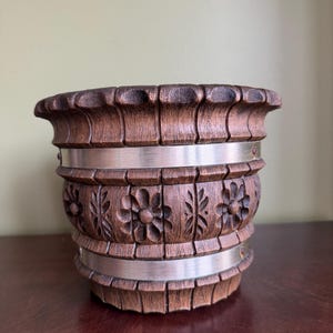 May include: A decorative wooden container with carved floral designs and two silver bands. The container has a dark brown finish and a fluted top and bottom. The container is on a dark wood surface.