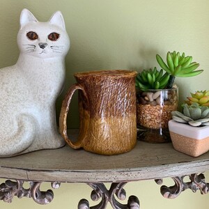 May include: A brown and tan ceramic mug with a textured surface sits on a shelf with a white ceramic cat figurine and potted succulents.