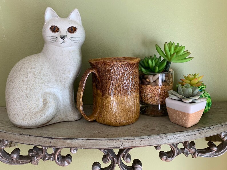 May include: A brown and tan ceramic mug with a textured surface sits on a shelf with a white ceramic cat figurine and potted succulents.