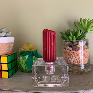 Puede incluir: Una vela roja en un portavelas de vidrio transparente, con una base cuadrada, se encuentra en un estante con tres pequeñas suculentas en macetas. Un cubo de Rubik colorido está en primer plano.