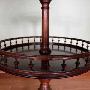 May include: A dark brown wooden two-tiered table with a round top and a decorative railing. The table has a central column and a pedestal base.