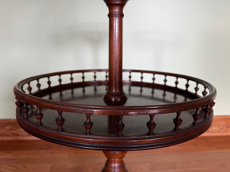 May include: A dark brown wooden two-tiered table with a round top and a decorative railing. The table has a central column and a pedestal base.