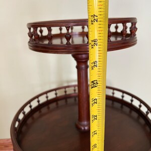 May include: A two-tiered wooden serving stand with a decorative railing around each tier. The stand is 27 inches tall and is made of dark wood.