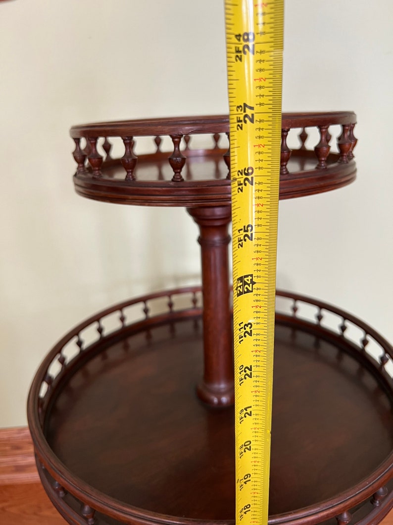 May include: A two-tiered wooden serving stand with a decorative railing around each tier. The stand is 27 inches tall and is made of dark wood.