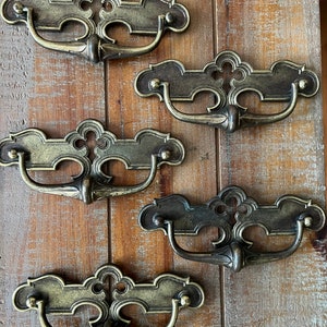 Könnte beinhalten: Fünf antike Messing-Schubladengriffe mit verzierten Designs. Jeder Griff hat einen geschwungenen, dekorativen Griff und eine rechteckige Platte mit einem erhabenen, komplizierten Muster.