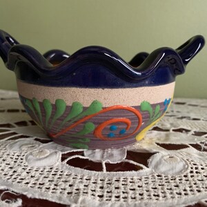 May include: A blue ceramic bowl with a scalloped rim and two handles. The bowl is decorated with a colorful floral design in shades of green, orange, yellow, and blue.