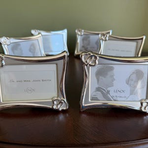 May include: A collection of four silver-toned metal picture frames with a curved rectangular shape. Each frame features a decorative double heart motif at the top left and bottom right corners. The Lenox brand logo is printed on the display inserts.