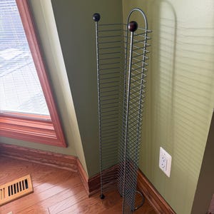 May include: A tall, freestanding media storage rack constructed from thin, grey metal wire. The unit features a vertical grid design with multiple slots for organizing discs and is topped with two dark brown, spherical wooden finials.
