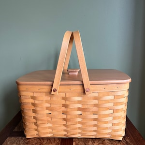 Puede incluir: Una cesta tejida natural con una tapa de madera y dos asas de madera. La cesta es de color marr&oacute;n claro y tiene un patr&oacute;n tejido. La cesta est&aacute; sentada sobre una superficie de madera.