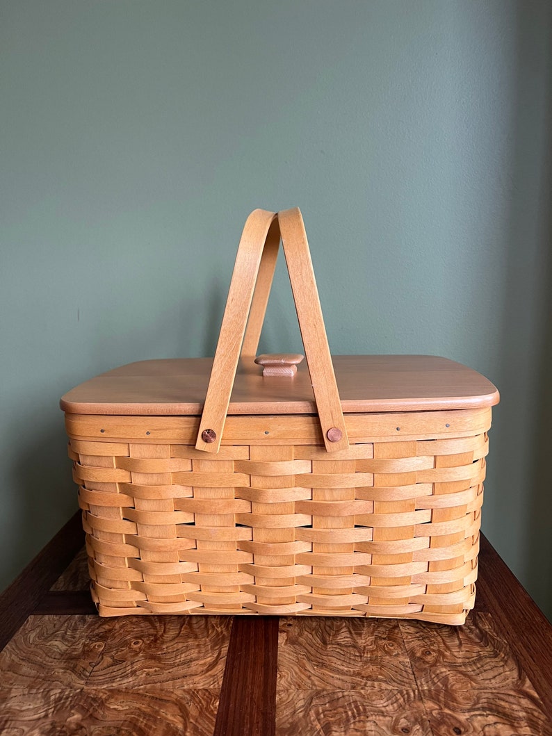 Puede incluir: Una cesta tejida natural con una tapa de madera y dos asas de madera. La cesta es de color marr&oacute;n claro y tiene un patr&oacute;n tejido. La cesta est&aacute; sentada sobre una superficie de madera.
