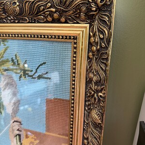 May include: A framed needlepoint artwork featuring a light blue background, a brown structure, and green foliage. The piece is encased in an ornate, heavy gold-toned frame with intricate relief carvings of floral and leaf motifs along the edges.