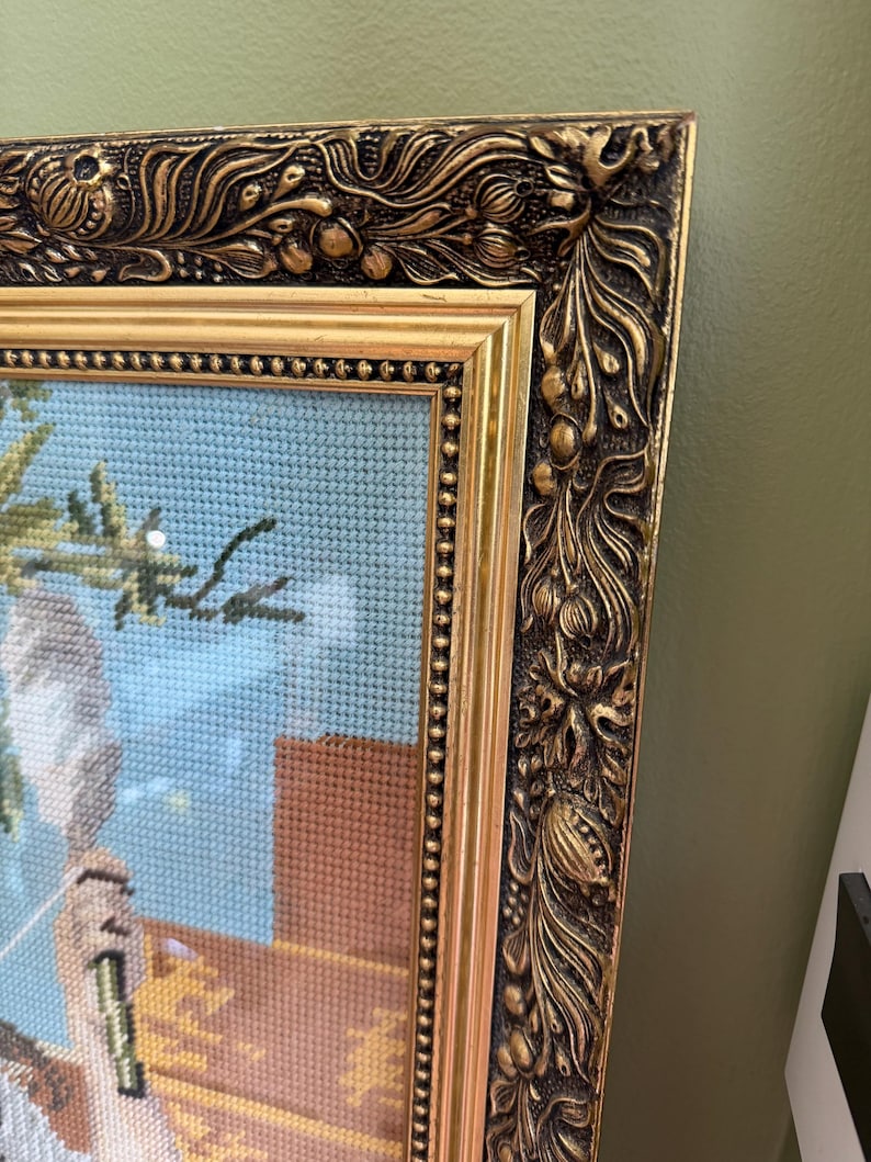 May include: A framed needlepoint artwork featuring a light blue background, a brown structure, and green foliage. The piece is encased in an ornate, heavy gold-toned frame with intricate relief carvings of floral and leaf motifs along the edges.