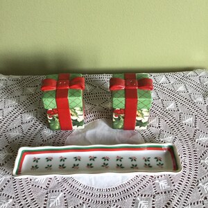 May include: Two green and red ceramic salt and pepper shakers shaped like wrapped presents with red bows. The shakers are on a white lace tablecloth with a white rectangular dish with a green and red border.