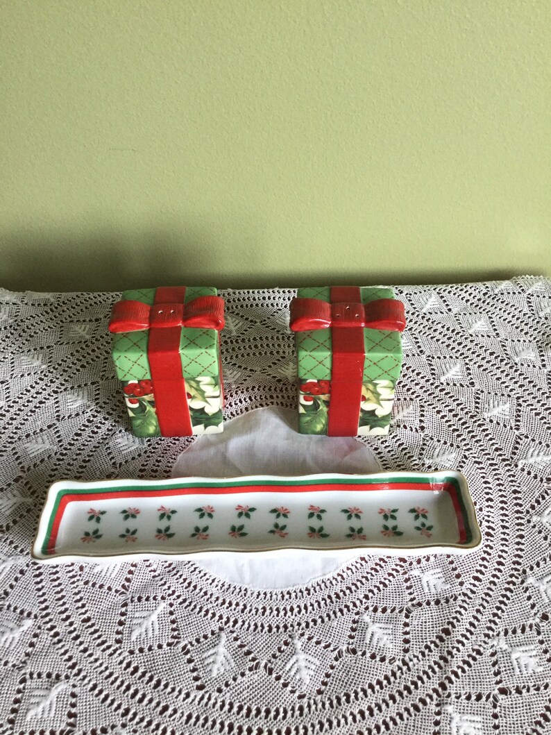 May include: Two green and red ceramic salt and pepper shakers shaped like wrapped presents with red bows. The shakers are on a white lace tablecloth with a white rectangular dish with a green and red border.