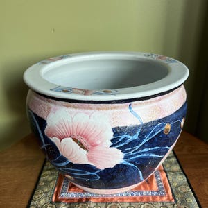 May include: A large, round, white ceramic planter with a blue and pink floral design. The planter has a wide rim and a slightly flared base. It is sitting on a small, square, patterned rug.