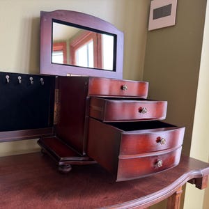 May include: A dark cherry-finished wooden jewelry armoire featuring a hinged lid with an interior mirror, a side-opening door with four metal hooks for hanging items, and three stacked pull-out drawers, each accented with a small floral knob.