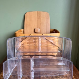 Puede incluir: Una cesta de picnic de madera con tapa y un juego de recipientes de pl&aacute;stico transparente divididos. Los recipientes est&aacute;n apilados uno encima del otro.