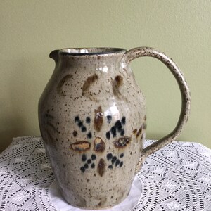 May include: A brown ceramic pitcher with a speckled glaze and a black and brown geometric design. The pitcher has a wide mouth and a handle.