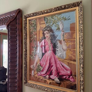 May include: A framed needlepoint tapestry depicting a person with long dark hair wearing a pink dress, sitting on a stone ledge while holding a spindle and thread. The artwork is encased in an ornate, textured gold-toned rectangular frame.