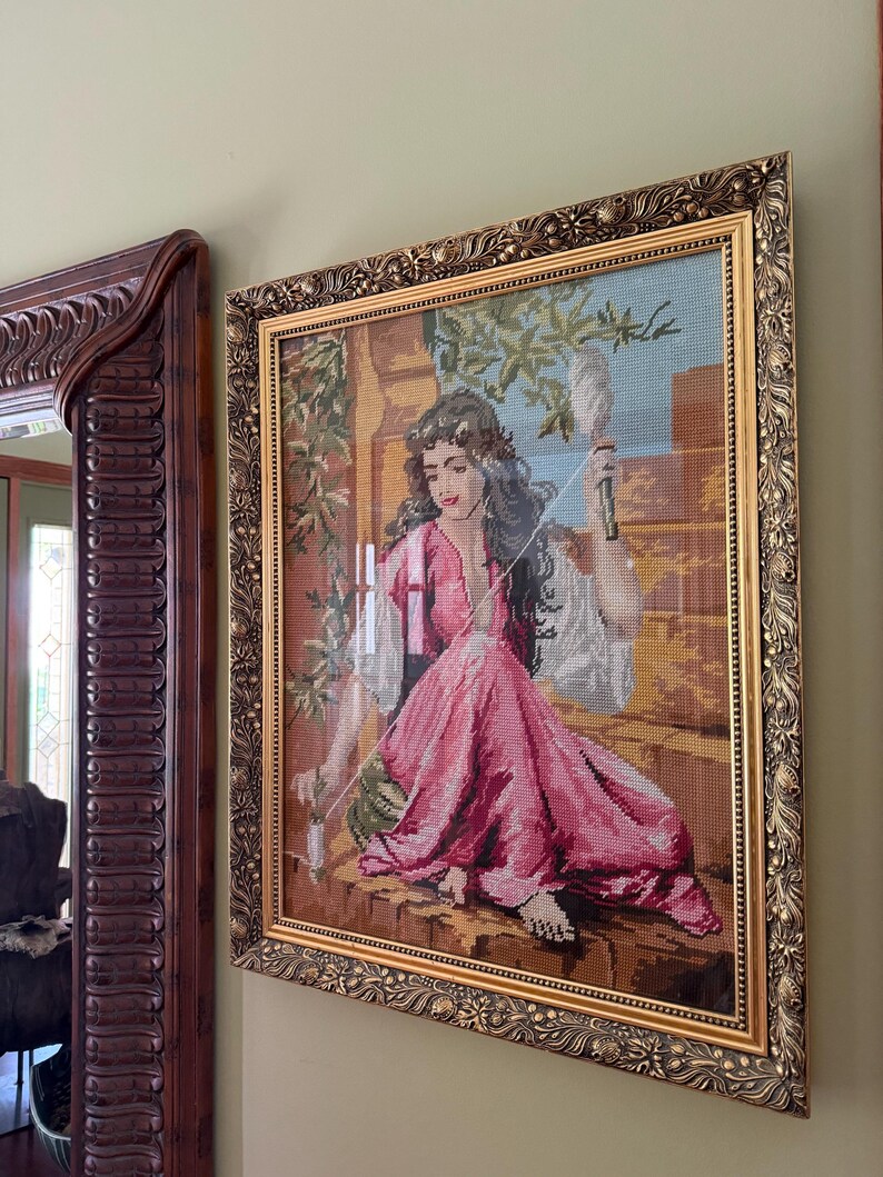May include: A framed needlepoint tapestry depicting a person with long dark hair wearing a pink dress, sitting on a stone ledge while holding a spindle and thread. The artwork is encased in an ornate, textured gold-toned rectangular frame.