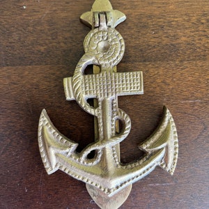 Puede incluir: Un llamador de puerta de ancla de latón con detalles intrincados. El ancla es un diseño náutico tradicional con un detalle de cuerda alrededor de la parte superior.