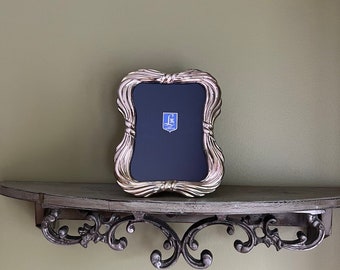 Puede incluir: Un marco de fotos plateado con un diseño ornamentado se encuentra sobre una repisa de madera con soportes de hierro decorativos. El marco tiene un paspartú negro y un pequeño emblema azul y dorado en el centro.