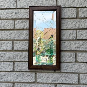 Puede incluir: Un espejo rectangular con marco marrón que refleja una escena de jardín con plantas verdes, una jaula para pájaros y muebles de exterior. El espejo está montado en una pared de ladrillos grises, creando un contraste.