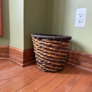 May include: A round, tapered wastebasket constructed from woven dark brown and natural tan reeds. The basket features a thick, braided rim at the top and a sturdy, flat base, showcasing a multi-tonal, textured pattern throughout its exterior surface.