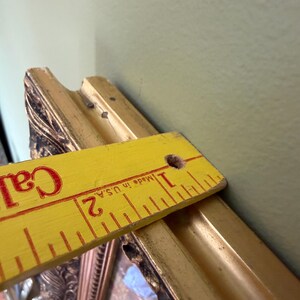 May include: An ornate gold-toned picture frame with a decorative relief pattern. A yellow wooden ruler is placed across the top edge, showing a frame depth of approximately 3.8 cm. The frame features a dark, weathered finish in the recessed areas.