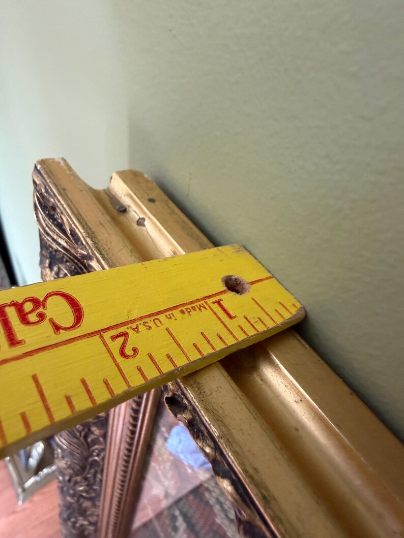 May include: An ornate gold-toned picture frame with a decorative relief pattern. A yellow wooden ruler is placed across the top edge, showing a frame depth of approximately 3.8 cm. The frame features a dark, weathered finish in the recessed areas.