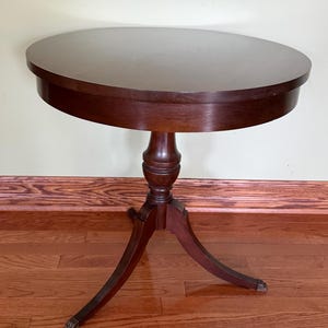 May include: A round, dark brown wooden pedestal table featuring a smooth circular top and a central column support. The base consists of three curved legs that extend outward, each ending in a small metallic foot detail at the bottom point.