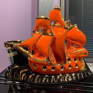 Puede incluir: Figurita de barco de cerámica naranja con detalles dorados. El barco está sentado sobre una base negra con detalles dorados. El barco tiene una vela y un mástil. La base tiene la palabra "Marsella" escrita en ella.