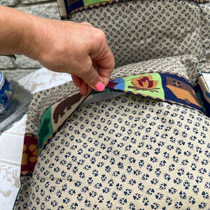 May include: A beige pillow with a blue paw print pattern. The pillow has a removable cover with a colorful design featuring a cabin, a campfire, a fish, and a bear.