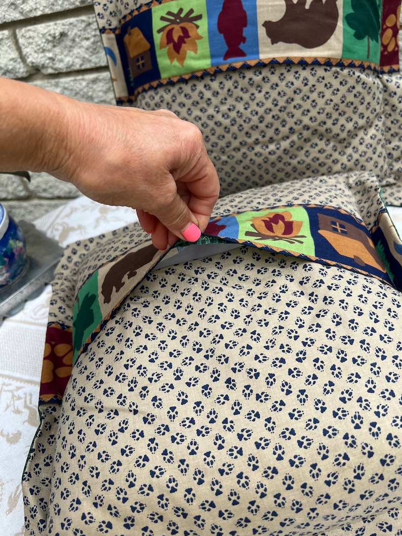 May include: A beige pillow with a blue paw print pattern. The pillow has a removable cover with a colorful design featuring a cabin, a campfire, a fish, and a bear.