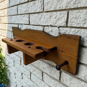 May include: A wall-mounted wooden rack featuring a horizontal shelf with three circular cutouts and a single protruding peg on the right side. The rack has a dark brown stained finish and is attached to the wall with two small metal chains.