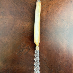 May include: A butter knife with a silver blade and a clear, textured glass handle. The blade has a serrated edge and a gold-colored accent near the handle. The handle is made of clear, faceted glass beads.