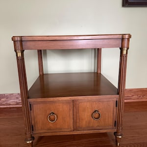 May include: A square wooden side table with a medium brown finish, featuring four fluted legs with brass-toned accents at the top and bottom. The table includes an open middle shelf and a lower cabinet section with two doors and brass ring pulls.