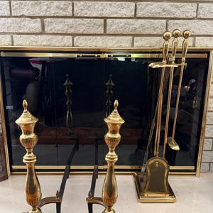 May include: A fireplace set featuring a black glass screen with gold trim, brass andirons, and a matching tool set. The andirons have a decorative, ornate design. The tool set includes a poker, shovel, and brush.