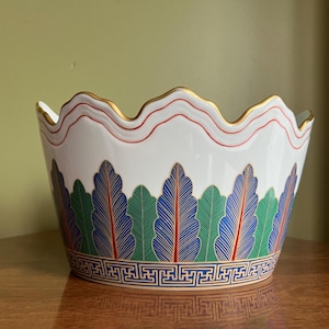 Puede incluir: Un cuenco de cerámica blanca con un borde festoneado y un borde dorado. El cuenco está decorado con un patrón de hojas verdes, azules y rojas y un borde geométrico dorado.