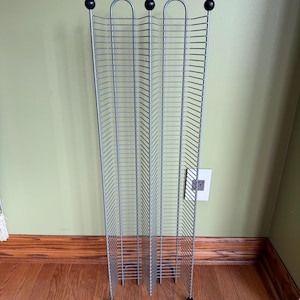 May include: A tall, silver-colored metal wire storage rack featuring a vertical design with multiple horizontal slots for organizing media cases. The structure has a zig-zag profile and is topped with three small, round black finials at the top.